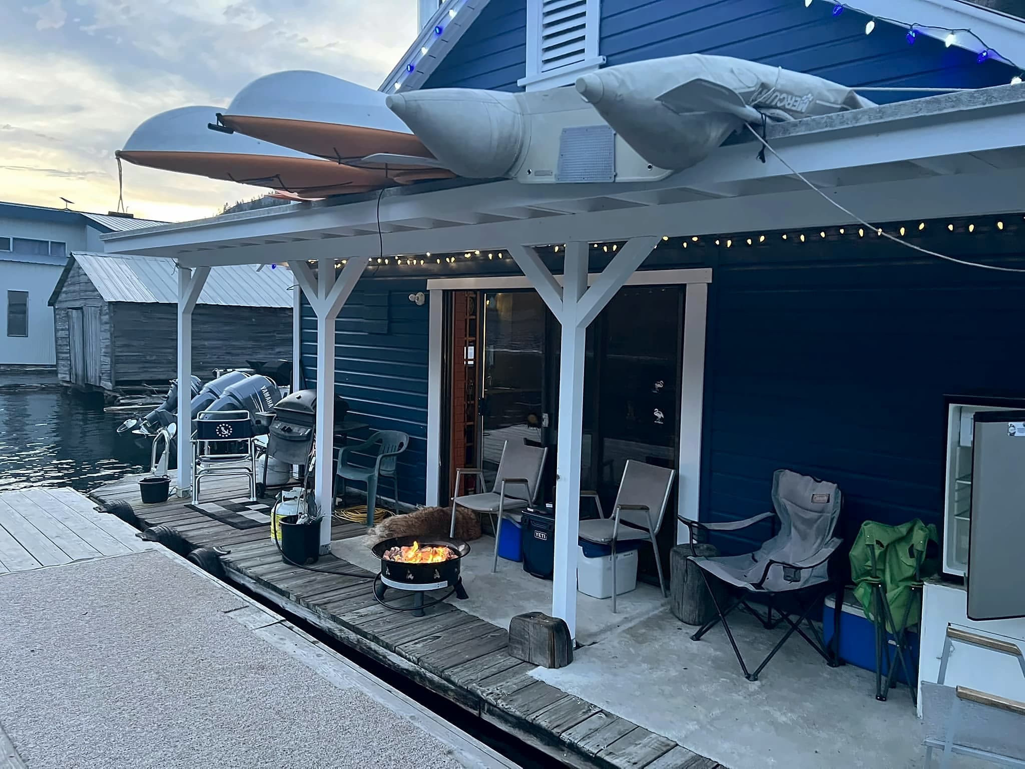 The floating cabin's deck at dusk: string lights over the chairs, a fire bowl lit, and kayaks stored on the roof.