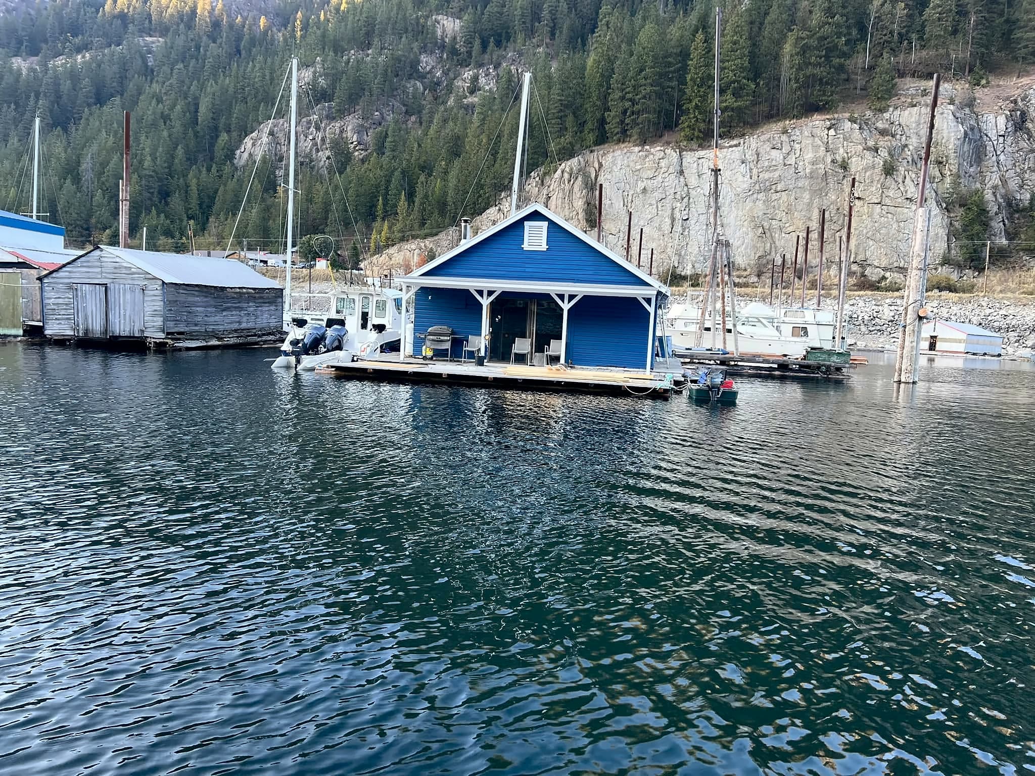 The blue floating cabin seen from the water, tied at the end of its own dock with the rock bluff behind it.