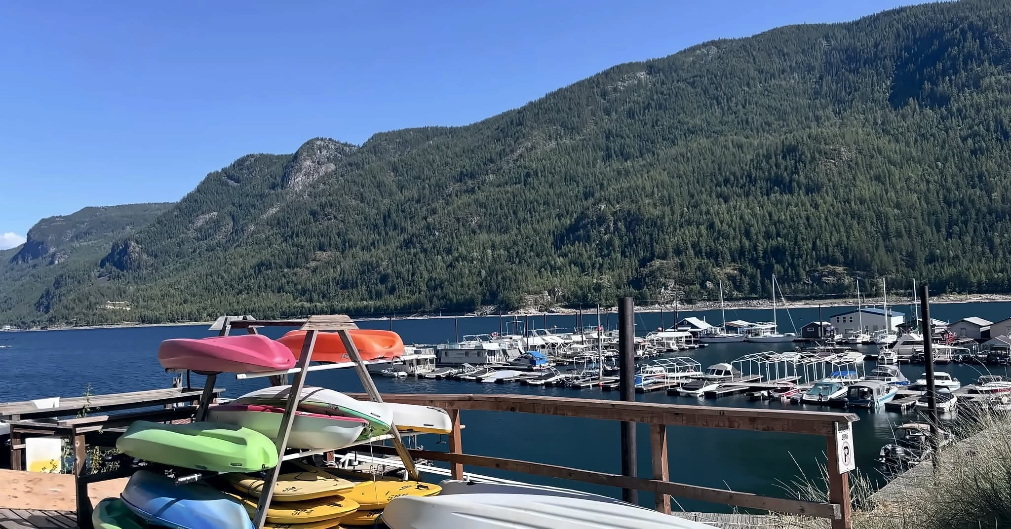 Kayaks and paddleboards stacked on a rack at the top of the dock, with the marina's boats and the forested mountains across the lake behind them.