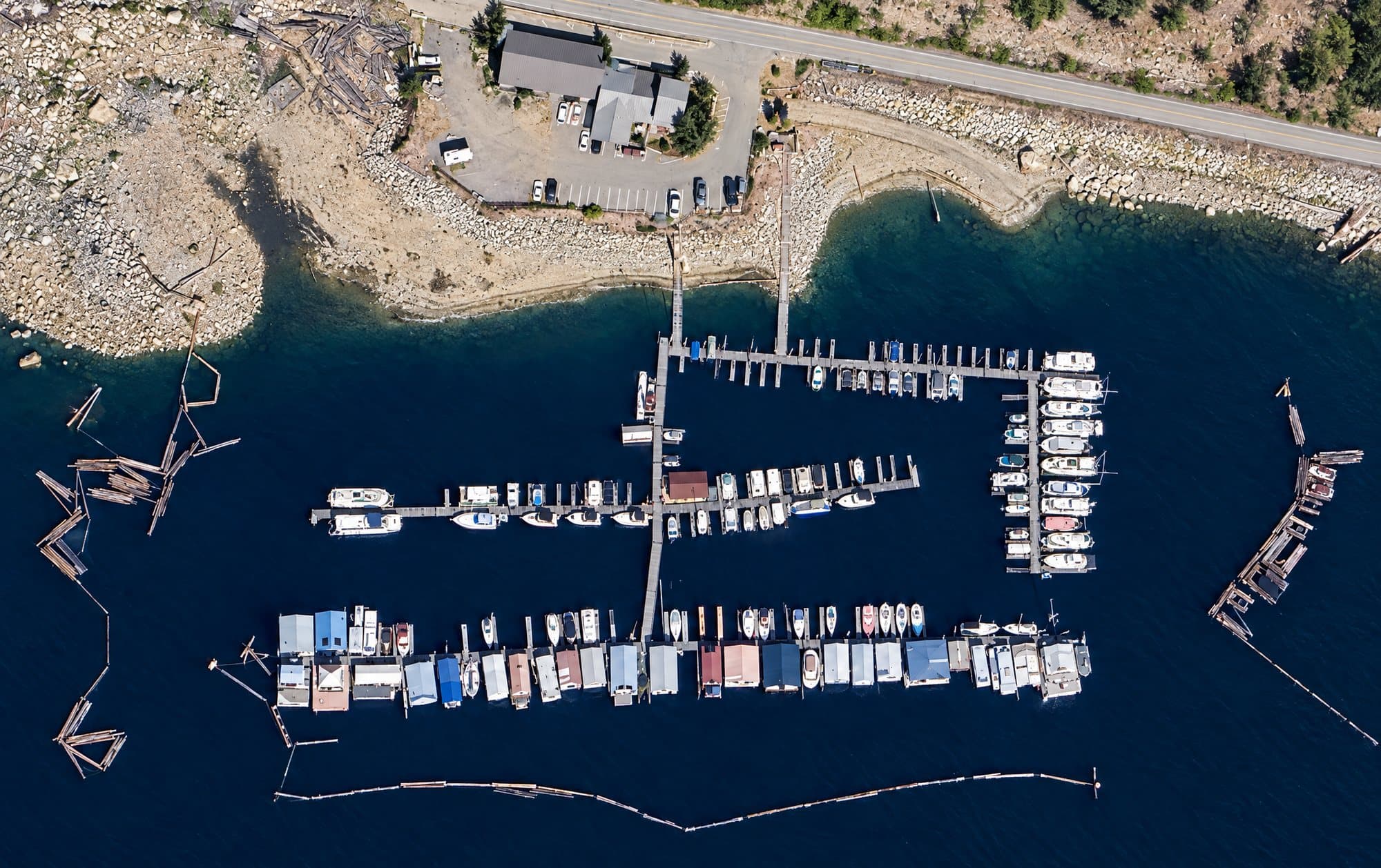 The marina from directly above: Broadwater Road and the buildings on the bank, the gangway down to the docks, three long docks of moorage and covered boathouses, a row of slips to the east, and the log booms lying off both sides.