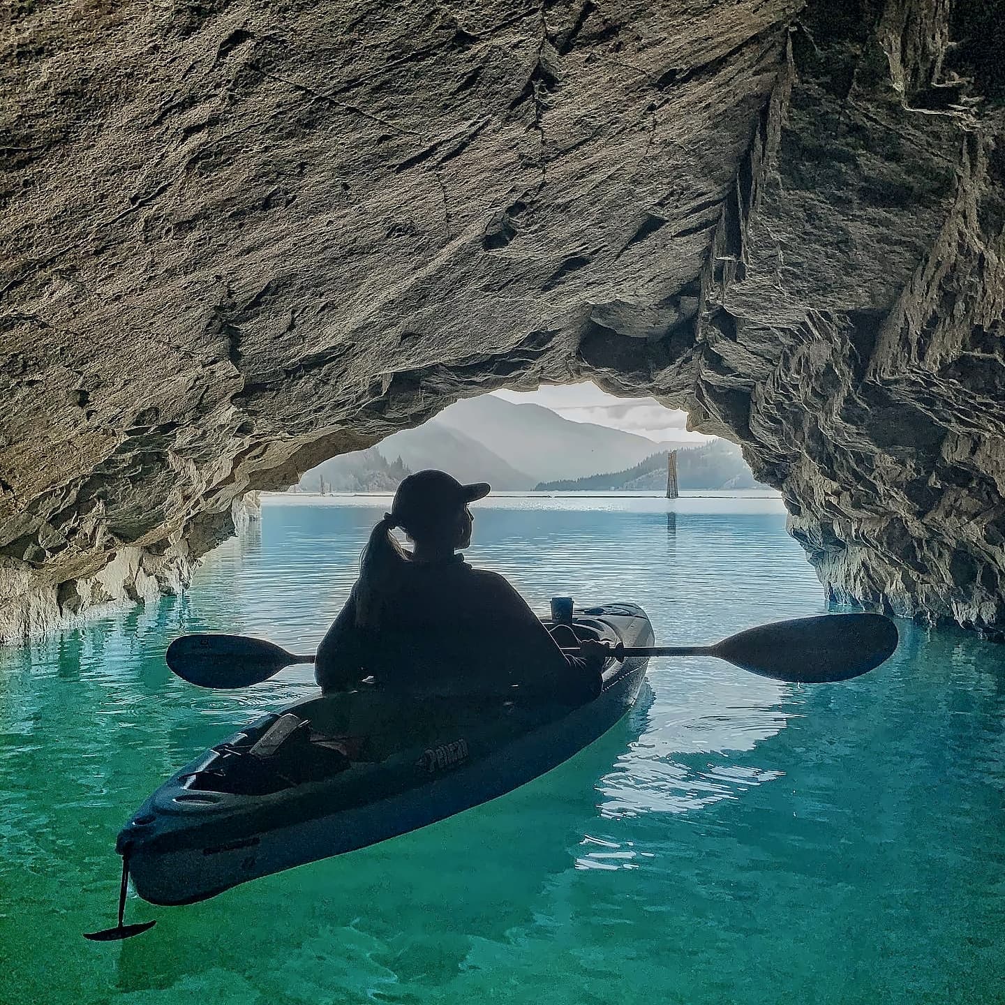 A paddler in a kayak inside the rock tunnel, looking out at turquoise water and the lake beyond.